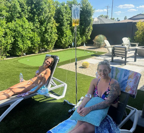 Two women in bikinis reclining on lounge chairs with an IV drip stand between them in a sunny desert-style backyard with putting green, cacti and patio seating.