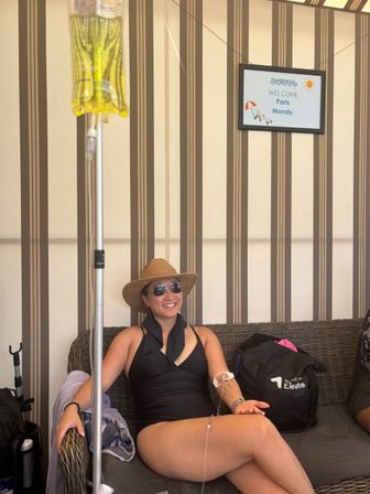 Smiling woman in a black swimsuit, sun hat and sunglasses relaxing on a wicker sofa inside a striped poolside cabana with an IV drip bag and tubing visible and a framed welcome sign on the wall — resort pool, summer vacation vibe.