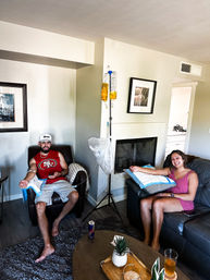 Two people lounging in a cozy living room, smiling as they receive at‑home IV hydration drips from yellow fluid bags hung on a stand, each with an arm IV, drinks and a small plant on the coffee table.