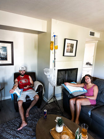 Two people lounging in a cozy living room, smiling as they receive at‑home IV hydration drips from yellow fluid bags hung on a stand, each with an arm IV, drinks and a small plant on the coffee table.