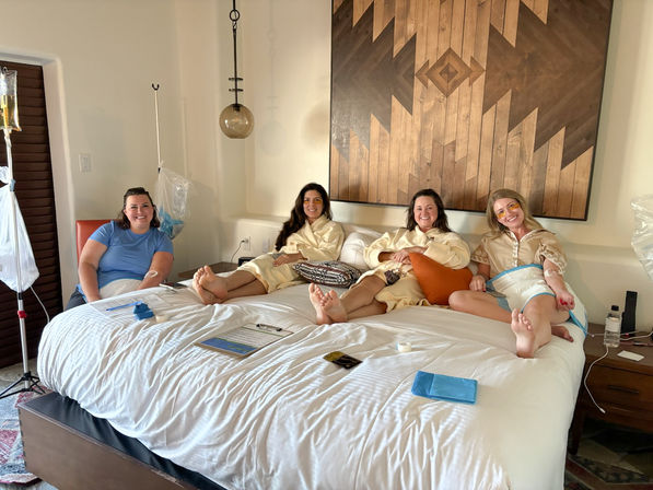 Four women lounging on a king-size bed in a bright wellness suite, wearing robes and under-eye patches and smiling while receiving IV drips from hanging infusion bags, with geometric wooden wall art behind them.
