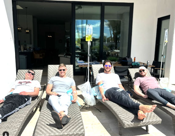 Four men in sunglasses relaxing on outdoor patio lounge chairs receiving IV drip therapy under sunny skies in front of sliding glass doors