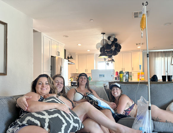 Four friends in swimsuits lounging on a gray couch in a bright open-plan living room and kitchen, smiling under a cluster of black-and-silver balloons with an IV drip pole and cooler nearby — relaxed at-home celebration.