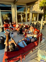 Group of people relaxing on a large red curved outdoor sectional on a sunlit modern patio in golden afternoon light, with IV drip stands, drinks, hanging chairs, patio heater and a palm-tree backed rock wall.
