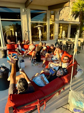 Group of people relaxing on a large red curved outdoor sectional on a sunlit modern patio in golden afternoon light, with IV drip stands, drinks, hanging chairs, patio heater and a palm-tree backed rock wall.