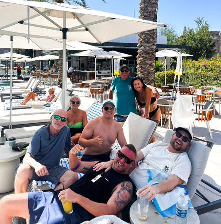 Cheerful group of friends lounging on poolside chairs at a sunny resort with palm trees, white umbrellas, cocktails, and an outdoor bar