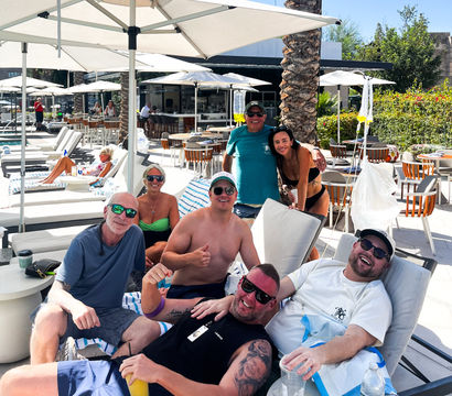Cheerful group of friends lounging on poolside chairs at a sunny resort with palm trees, white umbrellas, cocktails, and an outdoor bar