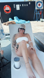 Smiling woman relaxing poolside on a white lounger in a white bandeau bikini and cap, sunglasses and tattoo sleeve, stainless tumbler in cup holder on a sunny outdoor pool deck with lifebuoy and pool rules sign on the wall.