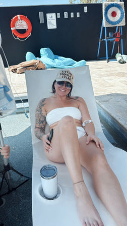 Smiling woman relaxing poolside on a white lounger in a white bandeau bikini and cap, sunglasses and tattoo sleeve, stainless tumbler in cup holder on a sunny outdoor pool deck with lifebuoy and pool rules sign on the wall.