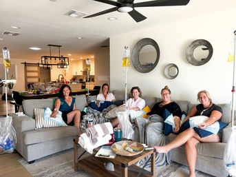 Five women seated on a sectional sofa in a bright, modern open-plan living room, each connected to IV drip bags for a group wellness hydration session, with blankets, pillows and a coffee table in the foreground and a kitchen in the background.