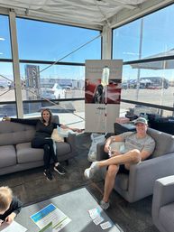 Two adults relaxing on gray sofas in a bright trackside motorsport hospitality lounge, man receiving an IV drip, race cars and paddock visible through large windows.