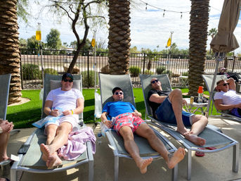 Four men lounging on poolside chairs under palm trees at a sunny desert resort-style outdoor area, wearing sunglasses and summer clothes, with IV hydration bags on poles and drinks on side tables.