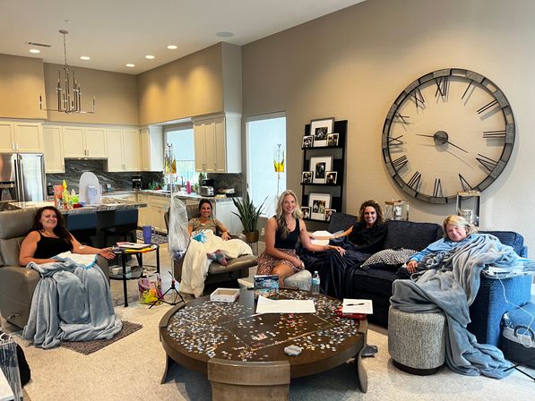 Five women relaxing under blankets in a modern open-plan living room with white kitchen cabinets, oversized wall clock, IV drip stands, and a jigsaw puzzle on the coffee table.