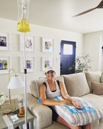 Smiling woman on a beige couch receiving a yellow IV drip at home — relaxed at-home hydration therapy in a bright living room with family photos and a coffee on the side table.