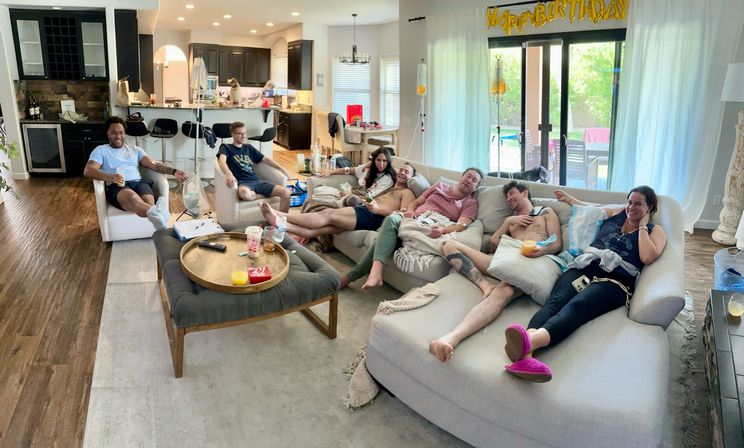 Group of friends lounging on a large sectional in a bright open-plan living room with a "Happy Birthday" banner, drinks on a coffee table, kitchen in the background and sliding glass doors to the backyard pool.