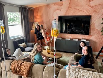 Group of smiling women enjoying an at-home IV hydration drip session in a cozy modern living room with a peach textured accent wall and wall-mounted TV