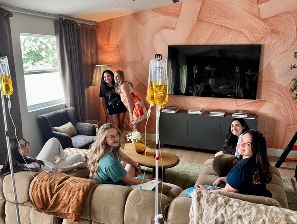 Group of smiling women enjoying an at-home IV hydration drip session in a cozy modern living room with a peach textured accent wall and wall-mounted TV