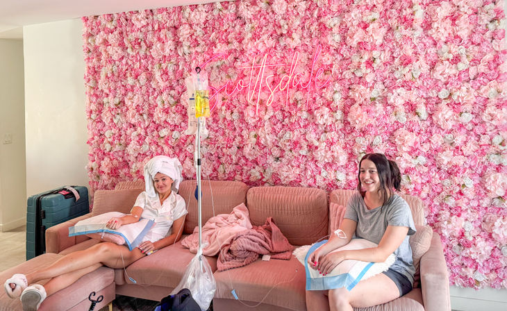 Two women relaxing on a pink couch receiving IV drips, smiling in a bright lounge with a pink floral wall and neon "Scottsdale" sign