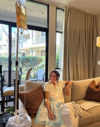 Smiling person relaxing on a couch with an IV drip in their arm, sunlit living room with floor-to-ceiling windows and a patio view, patterned throw pillows and a cozy blanket.