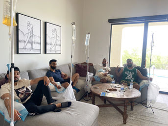 Four men relaxing on a sectional couch in a bright living room by a sliding glass door and pool, each hooked up to IV drip bags on stands during an at-home wellness session; wooden coffee table with water bottles and minimalist golfer wall art.