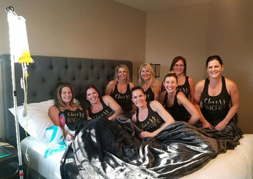 Eight women wearing matching "Cheers Bitches" tank tops smiling on a bed under a plush blanket with an IV drip stand beside them in a cozy bedroom, fun girls' night scene.