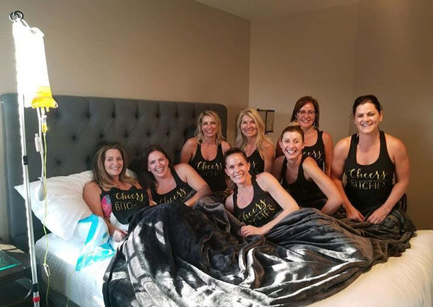 Eight women wearing matching "Cheers Bitches" tank tops smiling on a bed under a plush blanket with an IV drip stand beside them in a cozy bedroom, fun girls' night scene.