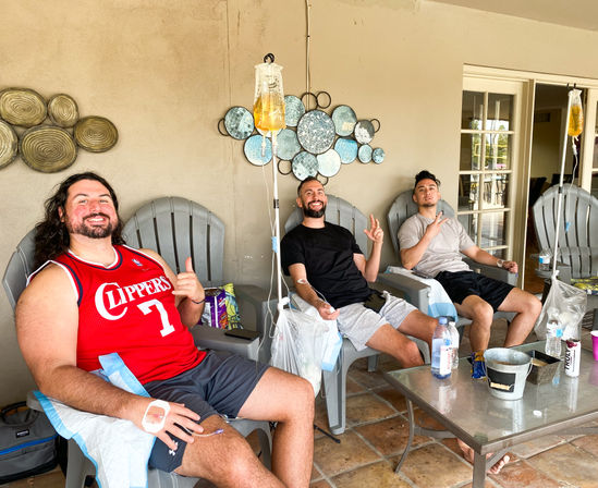 Three people relaxing on a covered residential patio in lounge chairs receiving IV hydration drips, smiling with water bottles and a small table of drinks and snacks.