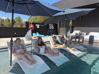 Four women in swimsuits lounging on submerged chaise lounges under navy umbrellas at a sunny rooftop pool lounge with IV hydration bags, a lifebuoy and poolside patio