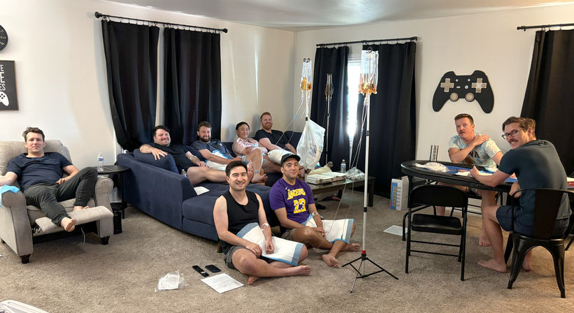 Group of men relaxing in a living room during an at-home IV hydration session, seated on couches, chairs and the floor with IV bags on stands and towels on their arms, gaming-themed decor on the wall.