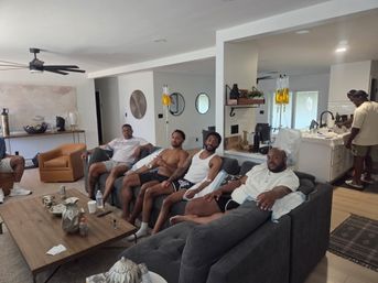 Group of men relaxing on a gray sectional in a modern open-plan living room with yellow IV hydration bags hanging near the kitchen, leather chairs and a wooden coffee table in view.