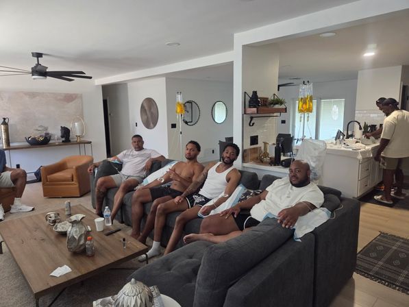 Group of men relaxing on a gray sectional in a modern open-plan living room with yellow IV hydration bags hanging near the kitchen, leather chairs and a wooden coffee table in view.