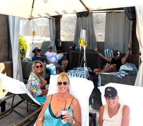 Group of smiling adults relaxing in an outdoor pool cabana on a sunny day, reclining on striped loungers with drinks and sunglasses while two IV hydration bags hang from poles, spa-style vacation scene.