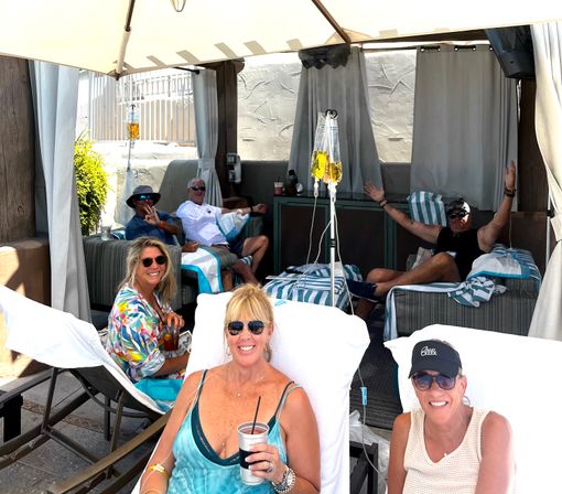 Group of smiling adults relaxing in an outdoor pool cabana on a sunny day, reclining on striped loungers with drinks and sunglasses while two IV hydration bags hang from poles, spa-style vacation scene.
