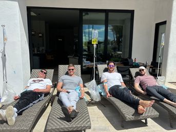 Four men relaxing on woven lounge chairs on a sunny residential patio, each hooked to IV hydration bags on stands by a sliding glass door — casual sunglasses, smiles, and a lighthearted outdoor wellness scene.