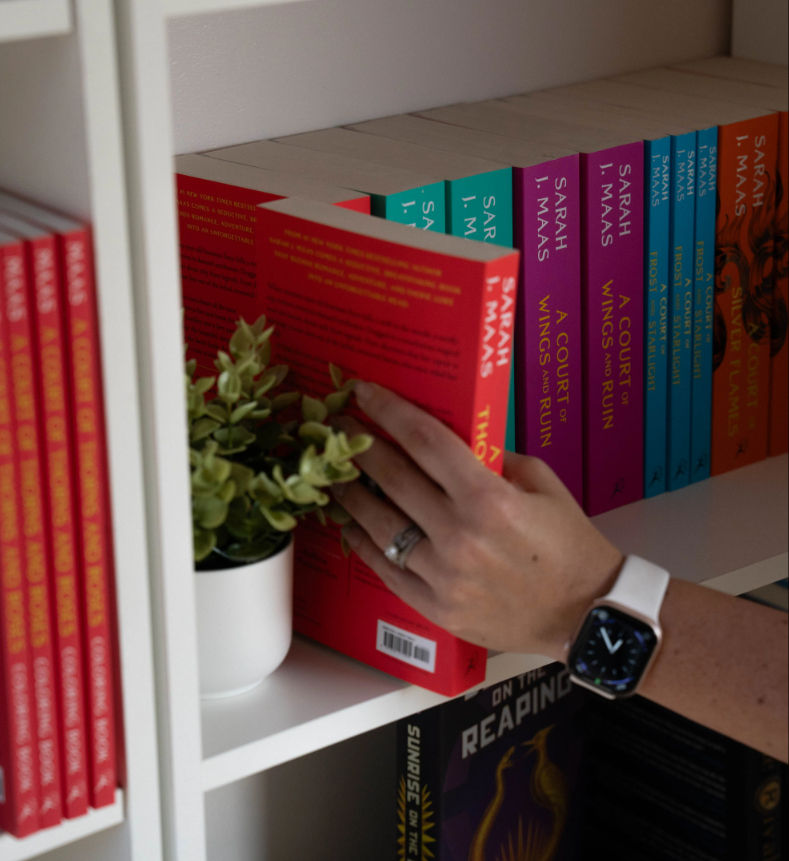 Hand reaching for a red paperback on a white home bookshelf filled with colorful fantasy paperbacks and a small potted succulent; wrist shows a smartwatch and a ring.