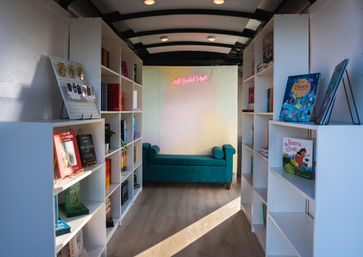Cozy bookmobile interior with white bookshelves stocked with colorful children’s and adult books, a teal chaise lounge at the back under a pink neon sign, and warm sunlight on wood-look floors.