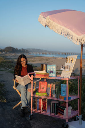 Woman reading on a coastal beach at golden hour beside a pink fringe umbrella and a pink wheeled cart stacked with colorful novels, with a pier and calm ocean in the background.