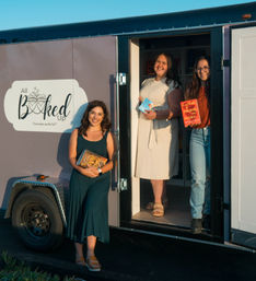 Three smiling women at the open door of a mobile bookstore trailer, each holding novels and enjoying warm golden-hour light.