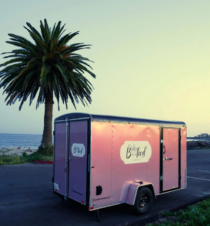 Pink cargo trailer parked in a beachside parking lot beside a tall palm tree with the ocean and pastel sunset sky in the background.