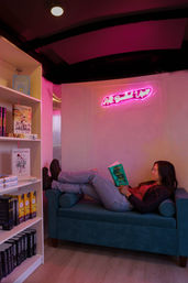 Person reclining on a teal loveseat in a cozy, pink-lit bookstore reading nook, holding 'A Court of Mist and Fury' beside a bookshelf under a neon script sign.
