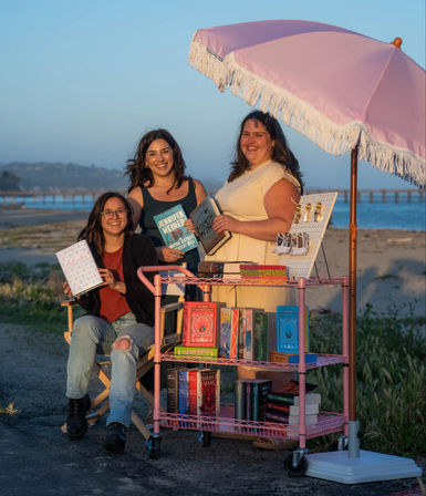 Beachside sunset pop-up book cart under a pink fringe umbrella — three smiling people holding books with colorful paperbacks and bookmarks on a rolling pink shelf.