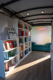 Sunlit cozy bookstore interior with white bookshelves of colorful books, teal upholstered bench, pink neon sign, arched ceiling and wood-look floor.