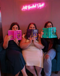 Three friends sit on a teal sofa in a pink neon-lit reading nook, each holding a colorful paperback that hides part of their faces — playful book club moment.