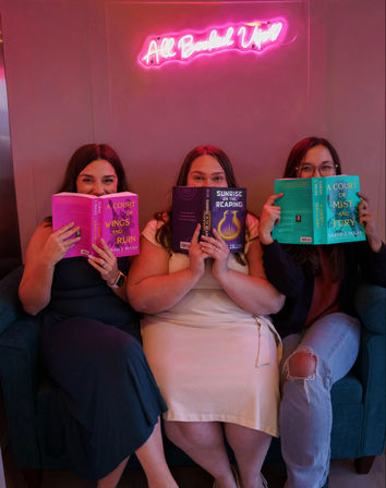 Three friends sit on a teal sofa in a pink neon-lit reading nook, each holding a colorful paperback that hides part of their faces — playful book club moment.