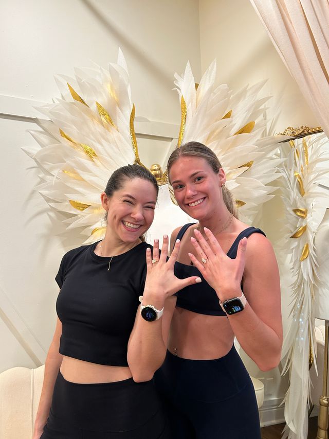 Two smiling women in black activewear showing rings and smartwatches in front of a decorative white-and-gold feather wing wall in a stylish studio