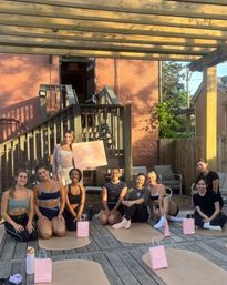 Smiling women in activewear gathered for an outdoor yoga class on a wooden deck under a pergola at golden hour, seated on mats with pink gift bags and an instructor holding a pink sign.