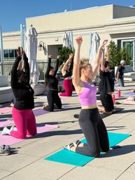 Sunny rooftop yoga class on an urban terrace — diverse participants kneeling on colorful mats with arms raised during an outdoor fitness/wellness session