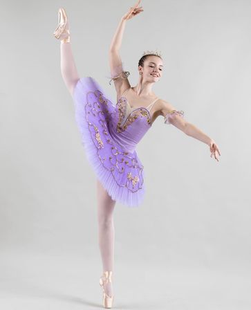 Classical ballet dancer in a lavender embellished tutu and tiara, balanced en pointe with a high leg extension and graceful arms against a neutral studio backdrop.
