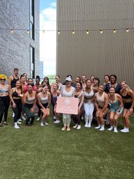 Outdoor urban rooftop group fitness class on artificial lawn between modern buildings, participants in activewear smiling under string lights and holding a pink sign.