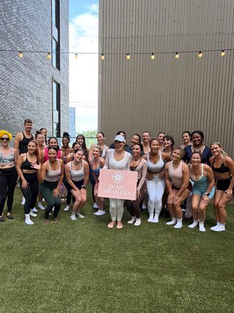 Outdoor urban rooftop group fitness class on artificial lawn between modern buildings, participants in activewear smiling under string lights and holding a pink sign.